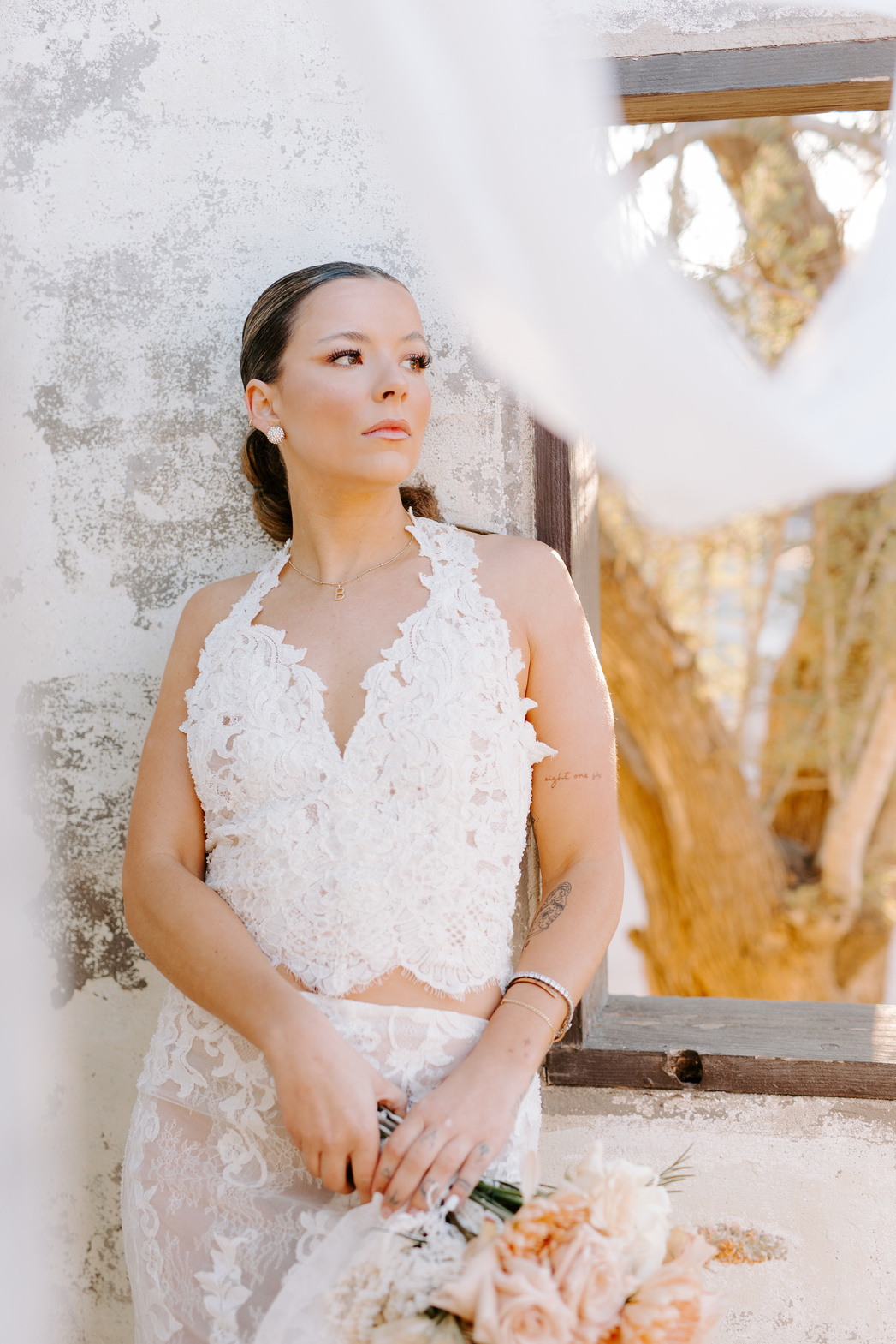 A beautiful bride in a white lace dress stands in front of a tree